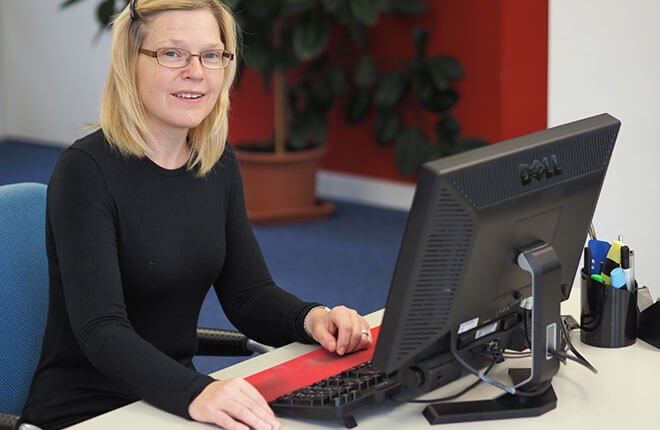 Heidi sitzt an einem Computerarbeitsplatz, die eingeschränkte rechte Hand liegt neben der Tastatur
